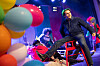 Scenekunstner ved en rød stol på en fargerik scene med ballonger og dansere.